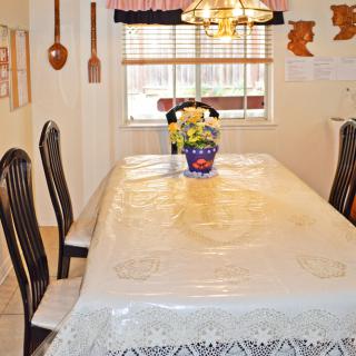 A dining room with a table covered in a lace tablecloth, surrounded by chairs and featuring a floral centerpiece under a chandelier.