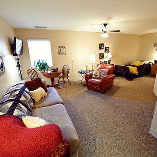 A cozy living room with sofas, armchairs, and a television, featuring warm decor and a carpeted floor.