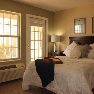 A cozy bedroom interior with a neatly made bed, soft lighting, and abundant natural light streaming through a large window.
