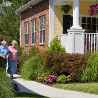 An elderly couple walks along a pathway beside a beautifully landscaped home with a welcoming front porch.