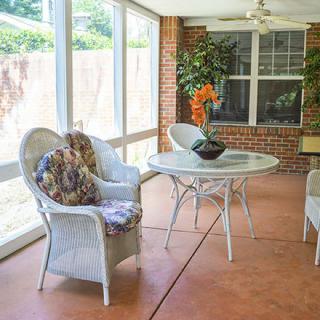 A bright sunroom with wicker chairs and a table, featuring an array of cushions and a potted plant by the window.