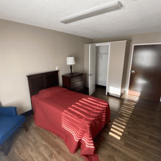 A minimalist bedroom featuring a single bed with a red blanket, a lamp on a bedside table, an open closet, and a wooden floor.