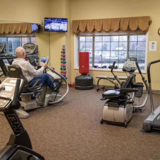 A senior person exercising in a well-equipped gym with various machines and a television.