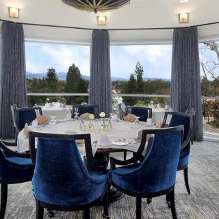 An elegantly decorated dining room featuring a round table surrounded by blue chairs, with large windows offering an outdoor view.