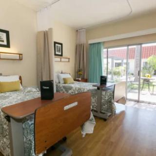 A hospital room with two patient beds, equipped with medical furniture and a view through large windows.