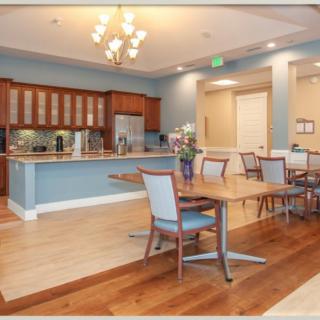 A modern dining area with a kitchen in the background, featuring wooden flooring, a chandelier, and neatly arranged tables and chairs.