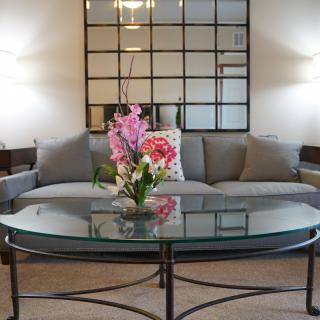 A modern living room with a grey sofa, glass coffee table, and decorative mirror, featuring a vibrant floral centerpiece.