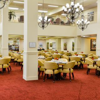 A spacious dining room with elegantly arranged tables and chairs, featuring chandeliers and a red carpeted floor.