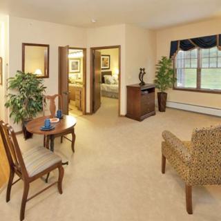 A cozy living room with chairs, a small table, plants, and a window allowing natural light.