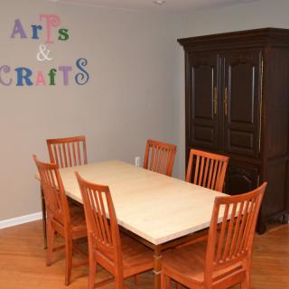 A dining room setup with a wooden table and chairs, featuring an 'Arts & Crafts' sign on the wall.