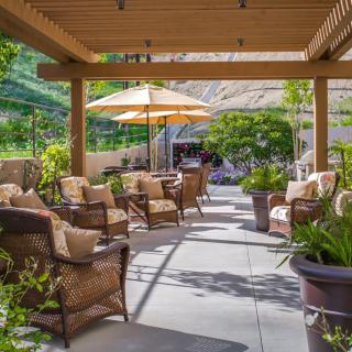A cozy patio with comfortable seating under a pergola, surrounded by greenery and shaded by an umbrella.