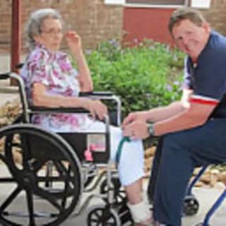 A caregiver tends to an elderly person in a wheelchair outside.