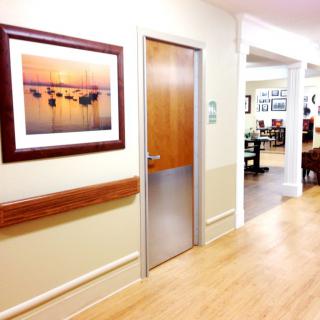 A brightly lit hallway with wooden flooring and a door, featuring framed artwork on the wall.