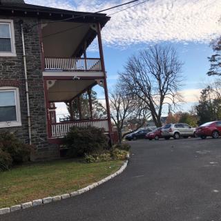 A residential building with a porch sits beside a parking lot filled with cars, set against a cloudy sky and surrounded by trees.