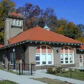 A historic brick building with a red tiled roof and large windows, surrounded by autumn trees.