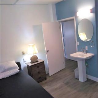A minimalist bedroom with a bed, nightstand, lamp, and adjacent sink area.