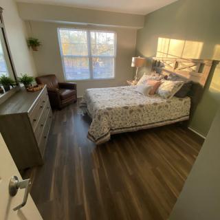 A cozy bedroom with a neatly made bed, dresser, and large window letting in natural light.