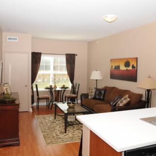 A cozy apartment living room with a sofa and dining area by the window, featuring warm tones and wood flooring.