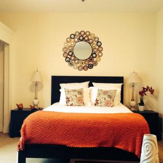 A stylish bedroom featuring a central bed with an orange blanket, decorative pillows, and symmetrical lamps. A decorative mirror serves as a focal point above the headboard.