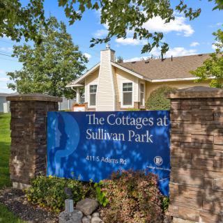 A sign for 'The Cottages at Sullivan Park' in front of a residential building surrounded by trees.
