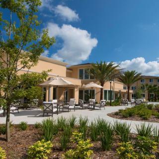 A modern building with outdoor seating surrounded by a well-maintained garden and palm trees under a blue sky with clouds.