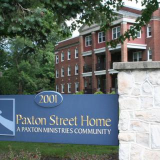 Sign for Paxton Street Home in front of a brick building surrounded by trees.