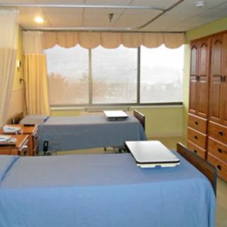 A hospital room with two beds, a large window, and wooden cabinets.