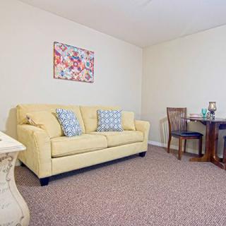 A cozy living room with a yellow sofa, decorative pillows, a small dining table with chairs, and artwork on the wall.