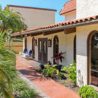 A charming Spanish-style house with arched windows and a terracotta roof, surrounded by lush landscaping and outdoor seating under a clear blue sky.