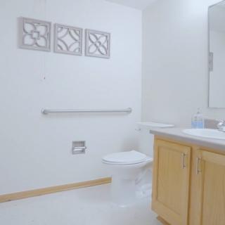 A clean and minimalist bathroom featuring a toilet, sink with a wooden cabinet, mirror, and decorative wall art.