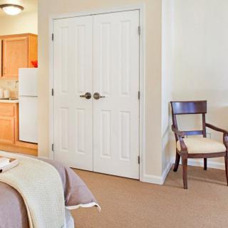A cozy bedroom with a view of the kitchen, featuring a chair, closet, and neatly made bed.