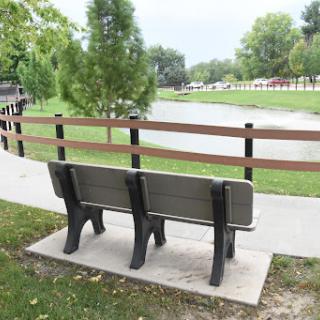 A peaceful view of a bench by a pond in a park, surrounded by trees and walking paths.