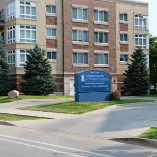 A multi-story brick building with a blue sign in front, surrounded by trees and road access.