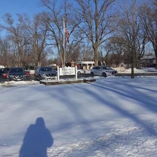 A serene winter landscape featuring a park blanketed in snow, with trees and parked cars under a clear blue sky.