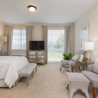 A spacious, neutrally decorated bedroom featuring a large bed, elegant furniture, and abundant natural light from large windows.
