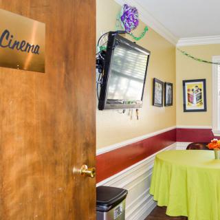 A cozy room interior featuring a wooden door labeled 'Cinema', with a wall-mounted TV and decorative elements above a round table covered with a green tablecloth.