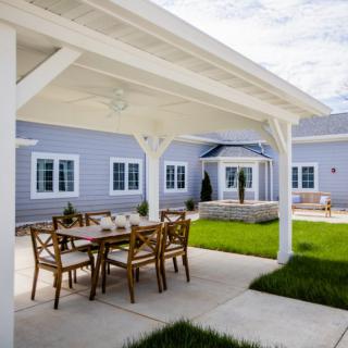 A covered patio with a dining table set in the backyard of a house, featuring a well-maintained lawn and landscaping.