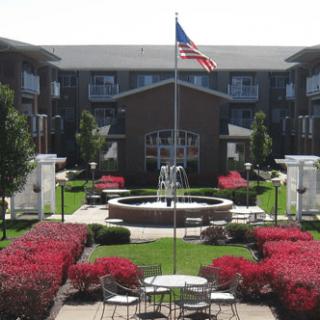 A well-maintained courtyard with a fountain, surrounded by an apartment building and featuring lush gardens and outdoor seating, with an American flag flying in the center.