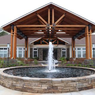 A beautifully designed building entrance with a central fountain, featuring wooden beams and landscaped surroundings.