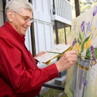 An elderly man joyfully paints a watercolor of flowers indoors.