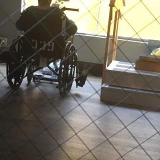 A person sitting in a wheelchair indoors, near a sunlit window and tiled floor.