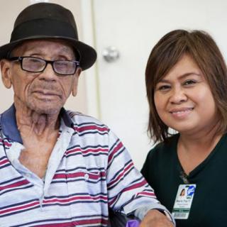 An elderly man wearing a hat and glasses sits next to a smiling woman, possibly a caregiver.