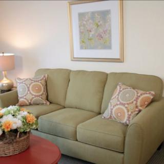 A cozy living room with a green sofa adorned with patterned pillows, accompanied by a side table with flowers and a lamp.