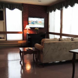 A cozy living room with a sofa and coffee table facing a television mounted on a brick wall, illuminated by a lamp next to large windows.