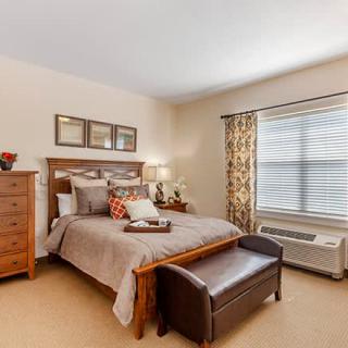 A cozy and elegantly decorated bedroom with a wooden bed frame, matching dresser, and a bench at the foot of the bed. The room is well-lit with natural light coming through a patterned curtain.