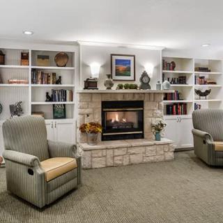 A cozy living room with a fireplace, armchairs, and bookshelves filled with decorative items.