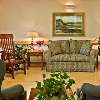 A cozy living room with a sofa, chairs, a table, and indoor plants, featuring a painting on the wall.