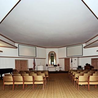 A spacious meeting room with rows of chairs, a pulpit, and flags in the background.