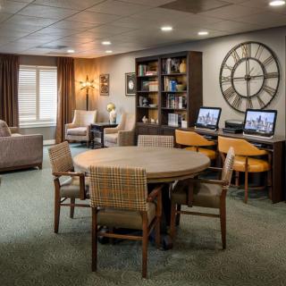 A cozy lounge area with a table and chairs, bookshelves, computers, and a large wall clock.
