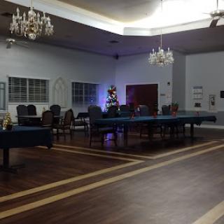 A spacious, elegantly decorated room featuring tables, empty chairs, and a Christmas tree, illuminated by chandeliers.